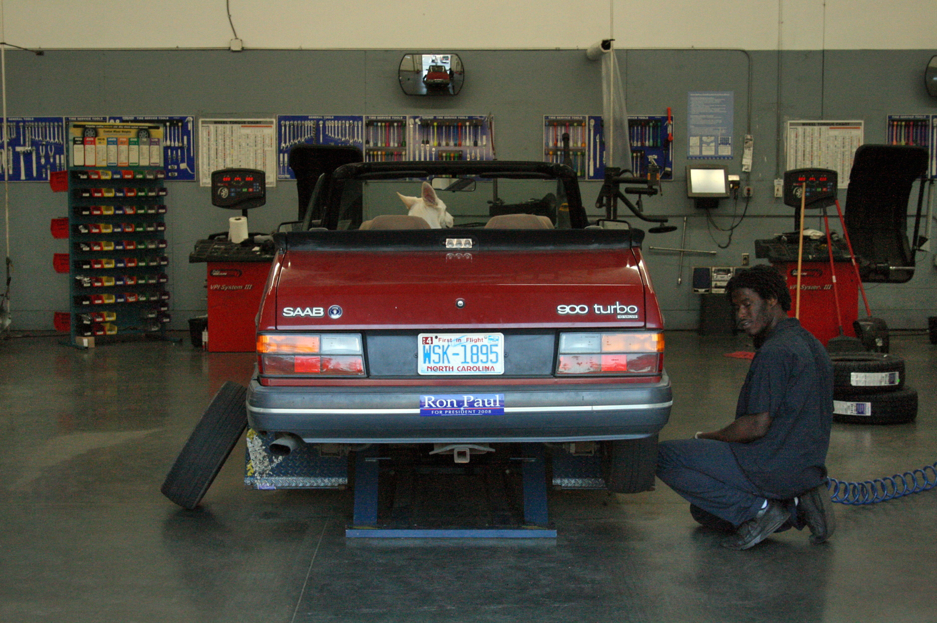 A red convertible raised on a hydraulic automotive service lift while a technician replaces the rear tires in a shop bay.