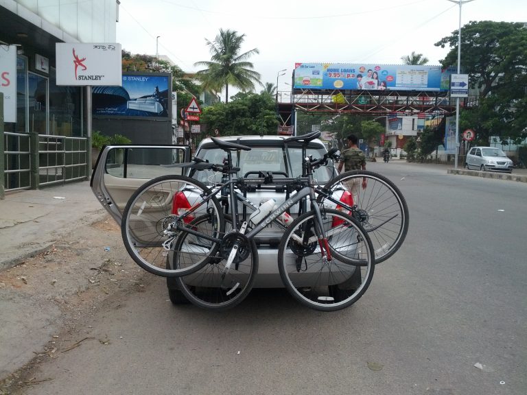 A trunk-mounted bicycle rack strapped to the rear hatch of a compact hatchback car, holding a bike in place with hook-and-strap arms.