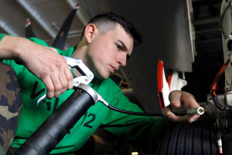 A mechanic uses a grease gun to lubricate a mechanical fitting during routine maintenance, the same type of lubrication point found on truck chassis and wheel bearings.