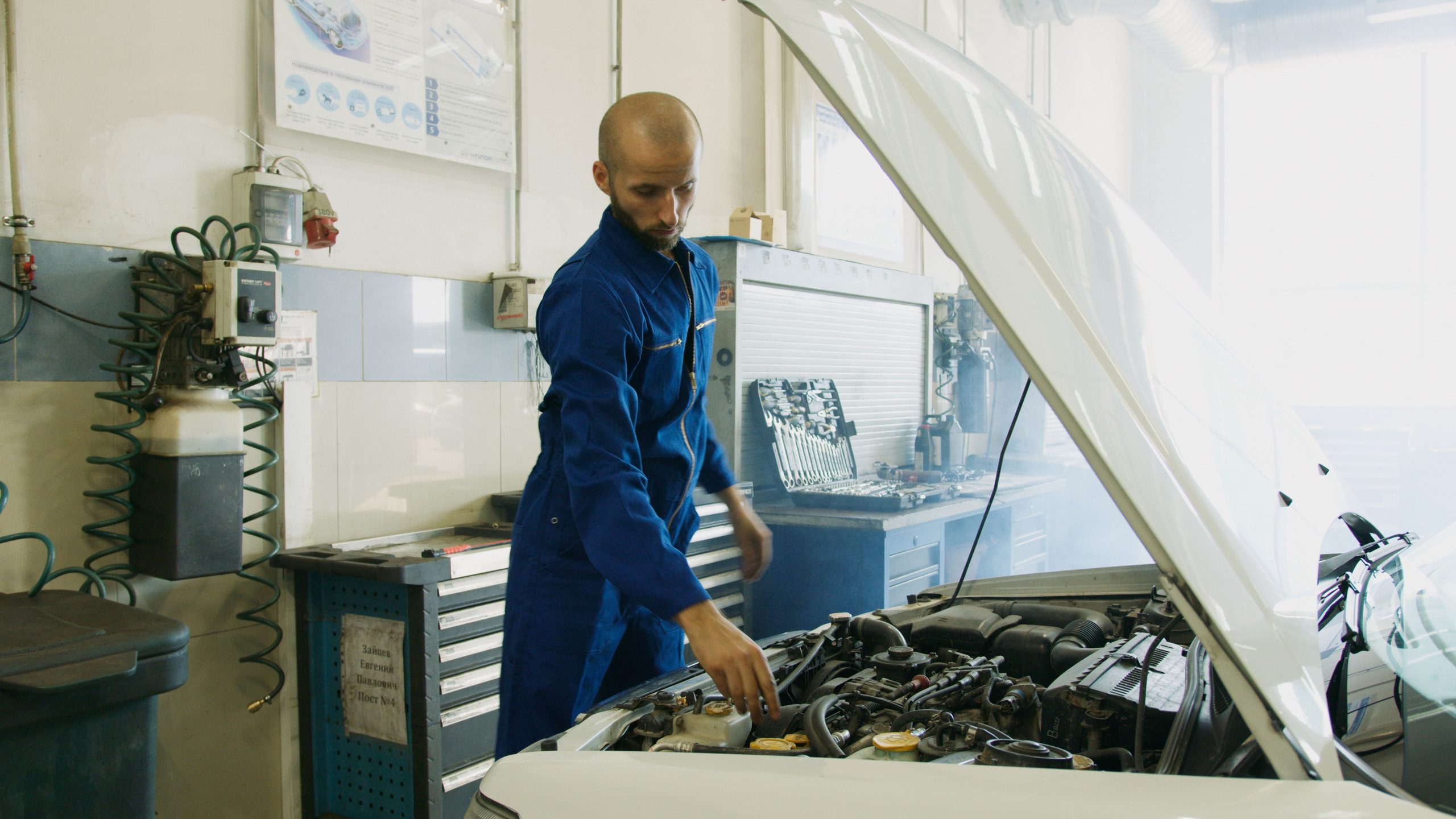 Mechanic in blue uniform leaning over and inspecting a car engine in a workshop.
